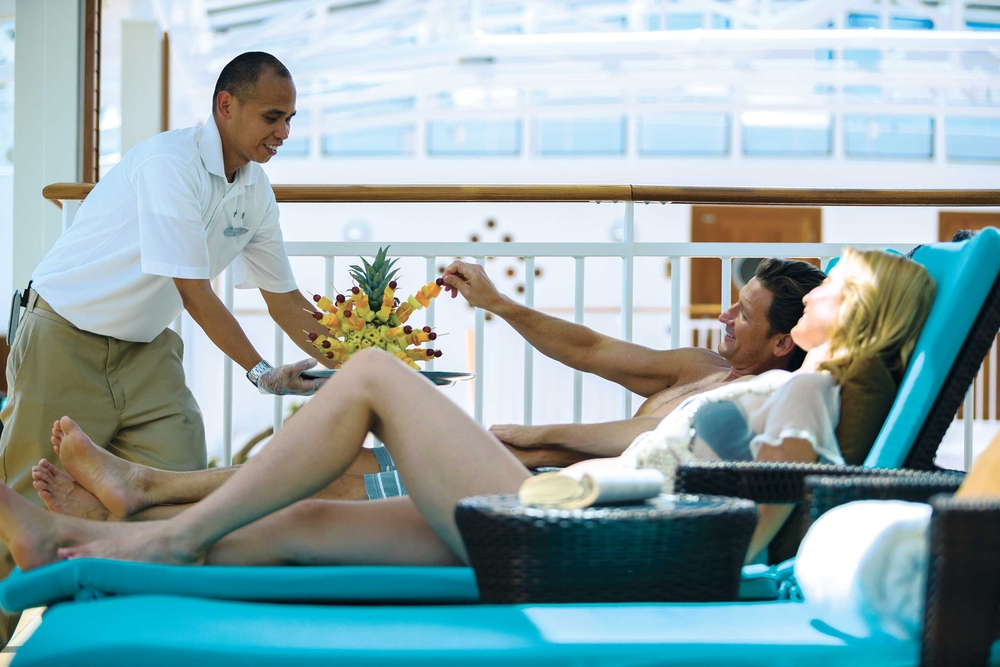 A man accepts fruit from a server while he and his wife relax in lounge chairs on deck.
