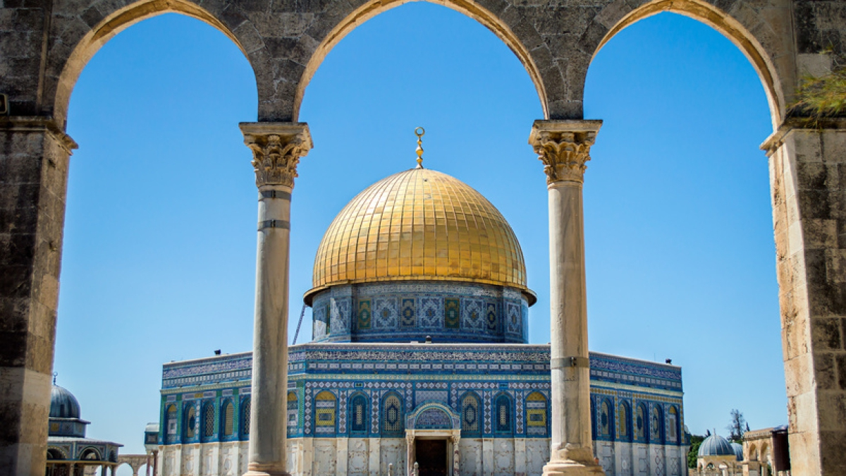 Photograph of thefamous Mosque (Dome of the Rock) in Jerusalem. The central building is seen through arched pillars, its blue tiles and gold dome shining in the sun.