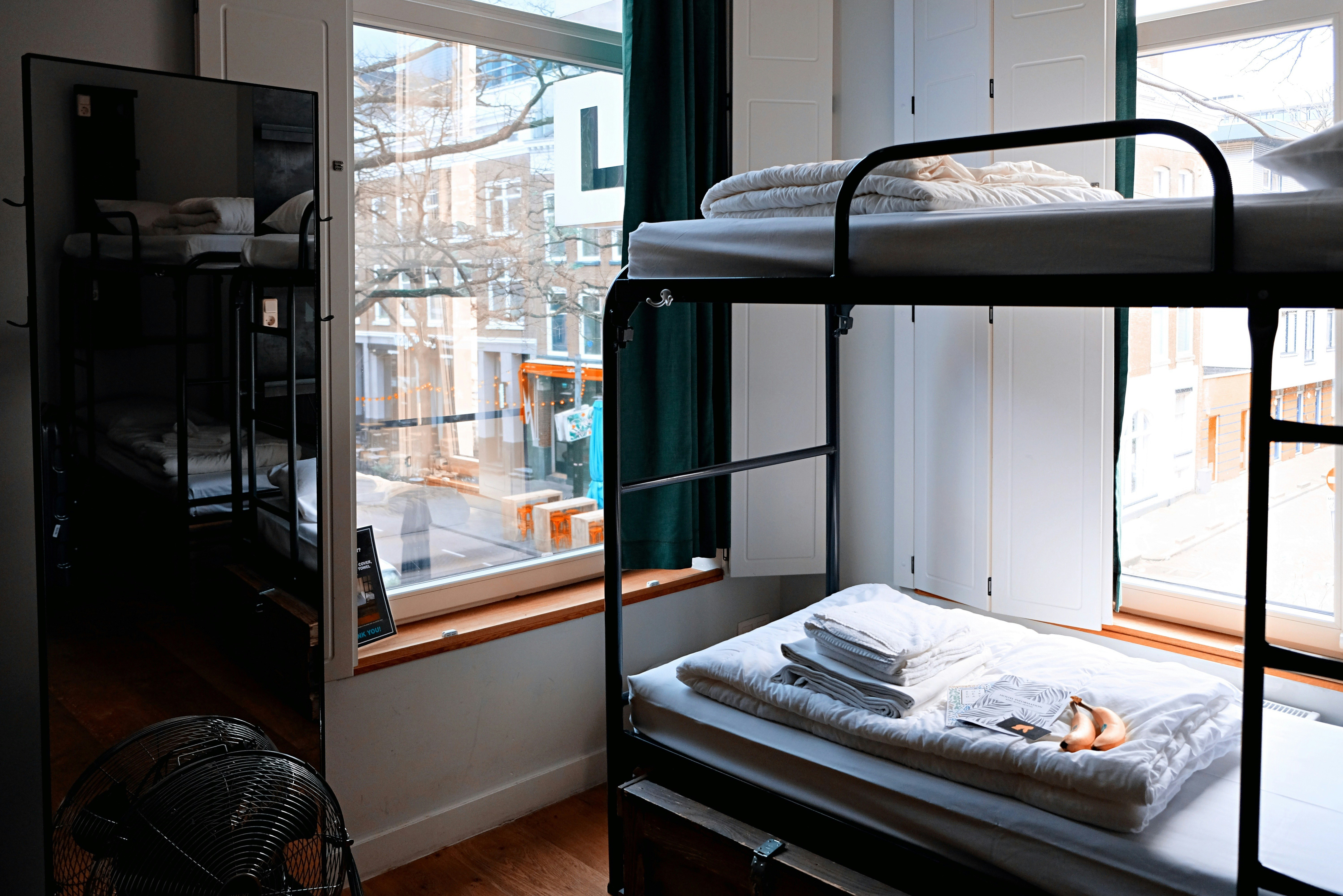 A bunk bed with clean folded linens is seen in this hostel room, with windows looking over a city.