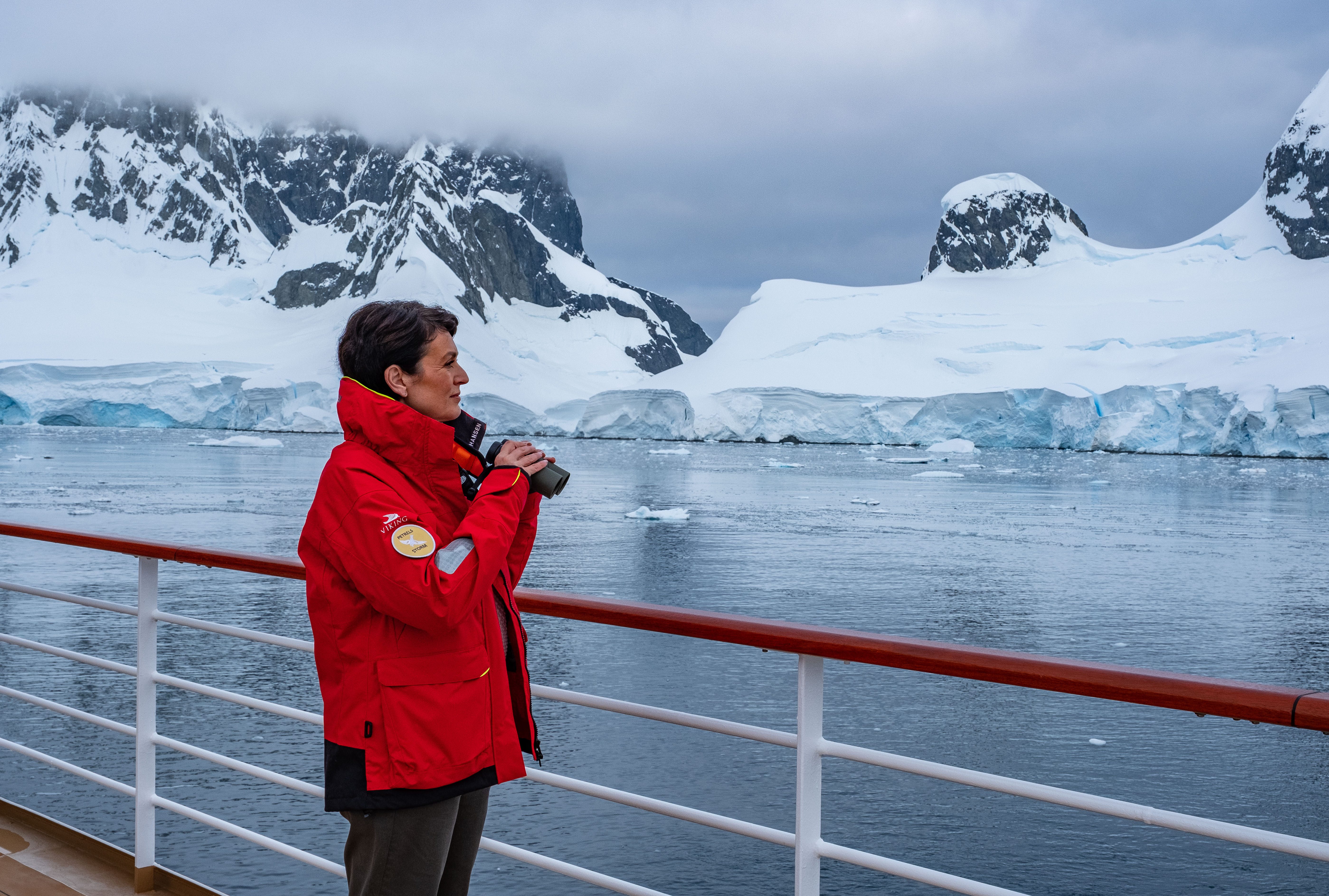 A woman wearing a red expedition coat looks out from a ship's rail with binoculars at the icy shores of Antarctica