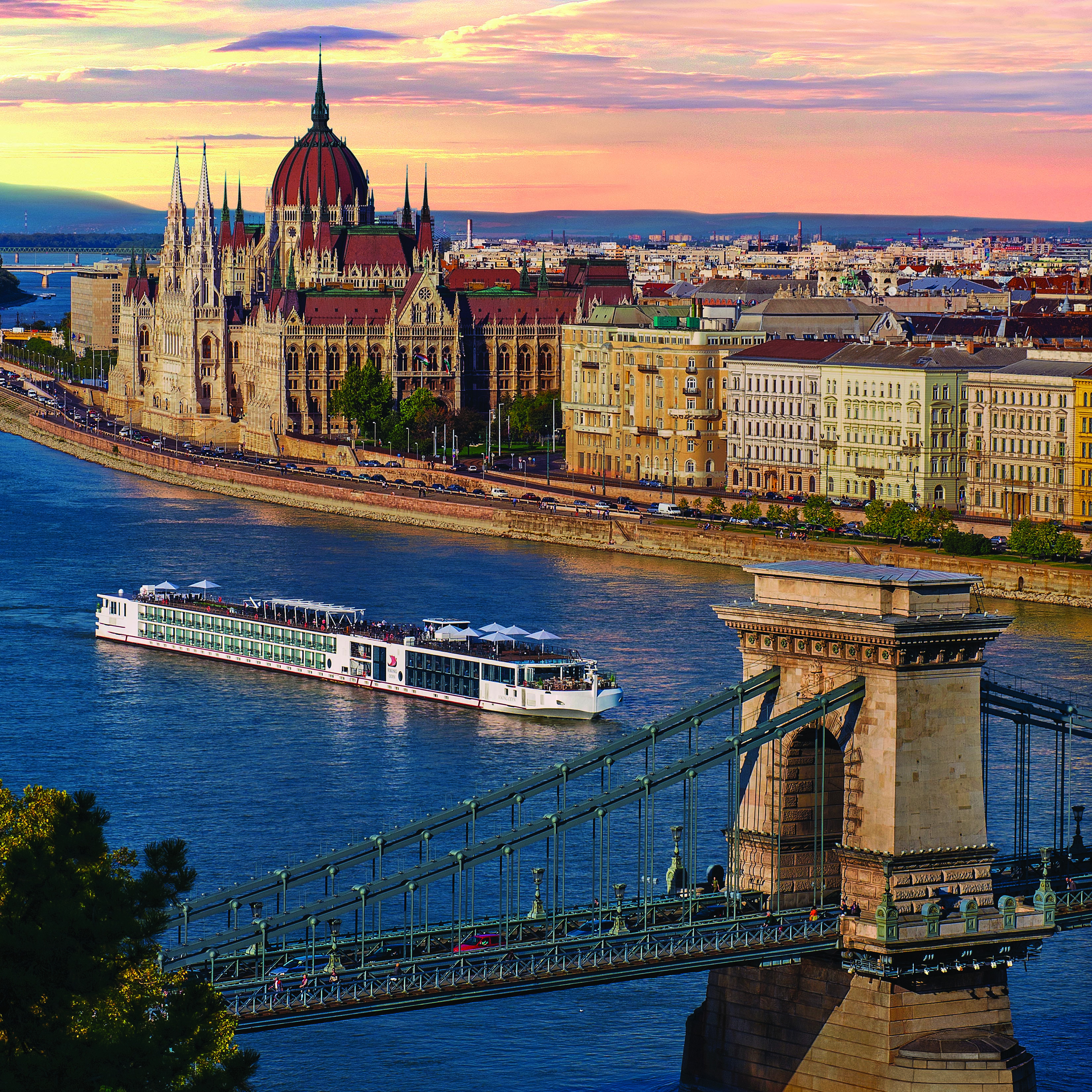 Photograph of the Chain Bridge in Budapest during a pink and yellow sunset, a river cruise is seen along the waterway