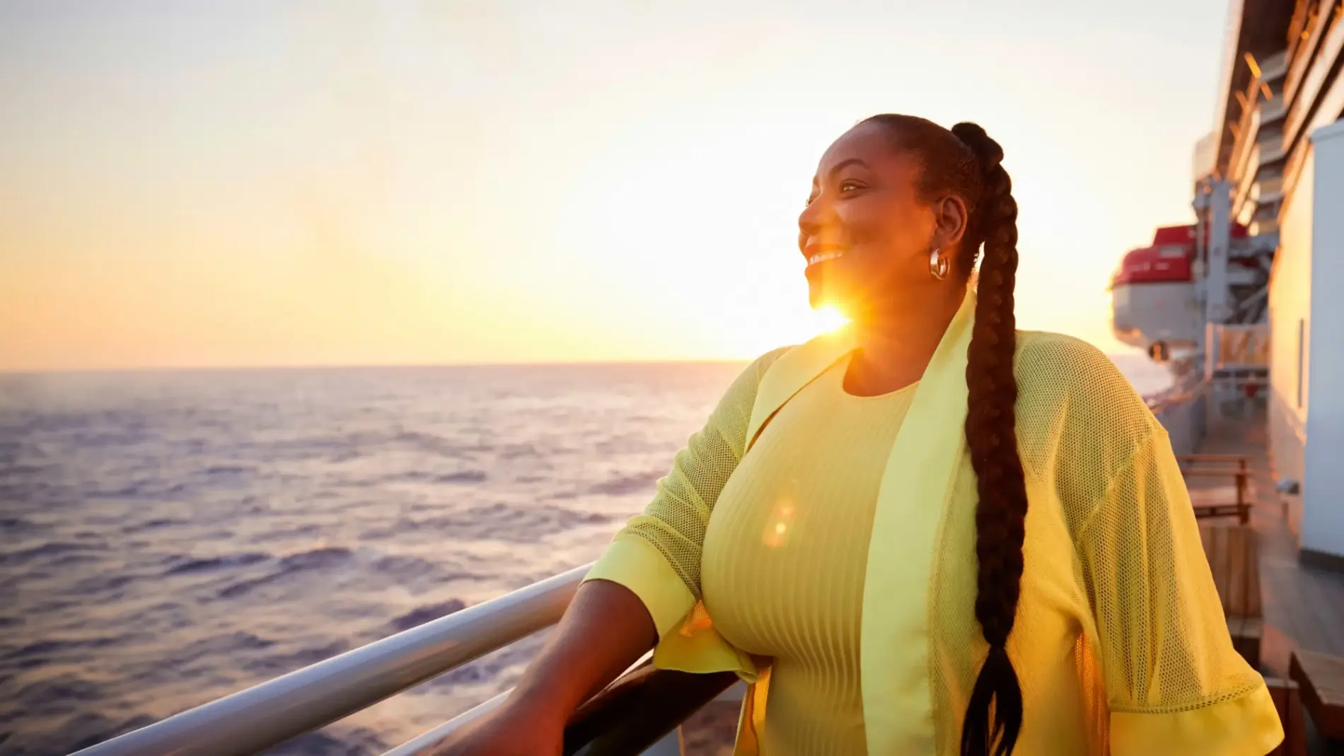 A dark skinned woman with long braided hair smiles in front of a sunset as she stands at the railing of a cruise ship.