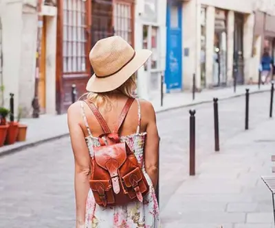 A woman explores a quaint european street wearing a sun hat, leather backpack, and sundress. The environment is summery and bright.