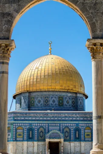Photograph of thefamous Mosque (Dome of the Rock) in Jerusalem. The central building is seen through arched pillars, its blue tiles and gold dome shining in the sun.