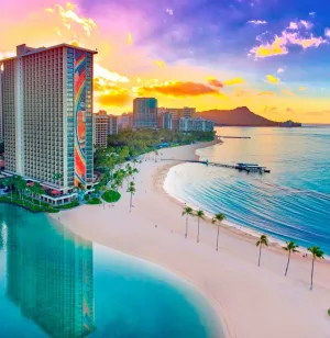 Photograph of the Hilton hotel towers along crystal blue waters and against a gorgeous yellow and purple sunset