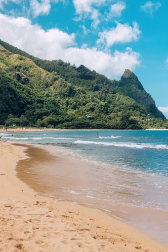 Photograph of sandy beaches in Kuai, a lush green mountain in the distance and calm blue waves nearby