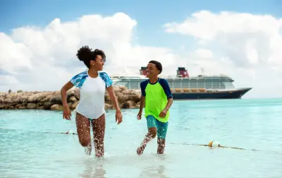Two children splash in shallow crystal blue waters on a tropical shore. A Disney Cruise vessel is visible in the background