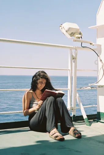A long haired woman reads a book while sitting against the railing on a cruise ship.