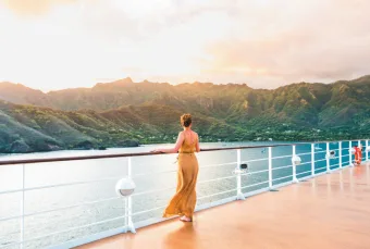 A woman looks off towards shore on the deck of a cruise ship. The weather is warm, and the sun sets over gorgeous scenery