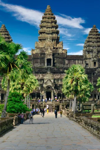 Photograph of Angkor Wat in Cambodia, taken by photographer Paul Szewczyk. Tourists walk along groomed green exteriors leading up to the ancient stone buildings